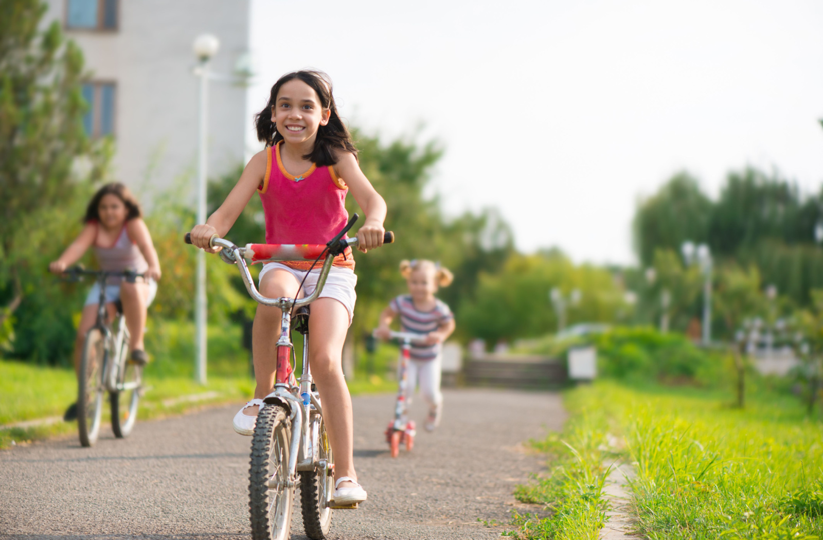 3 young girls smiling and riding their bikes in a park on a warm day.
