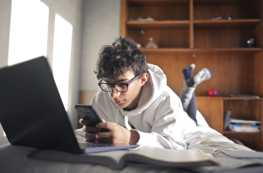 Young boy in a white sweater wearing glasses studying for school using his laptop and phone at the same time